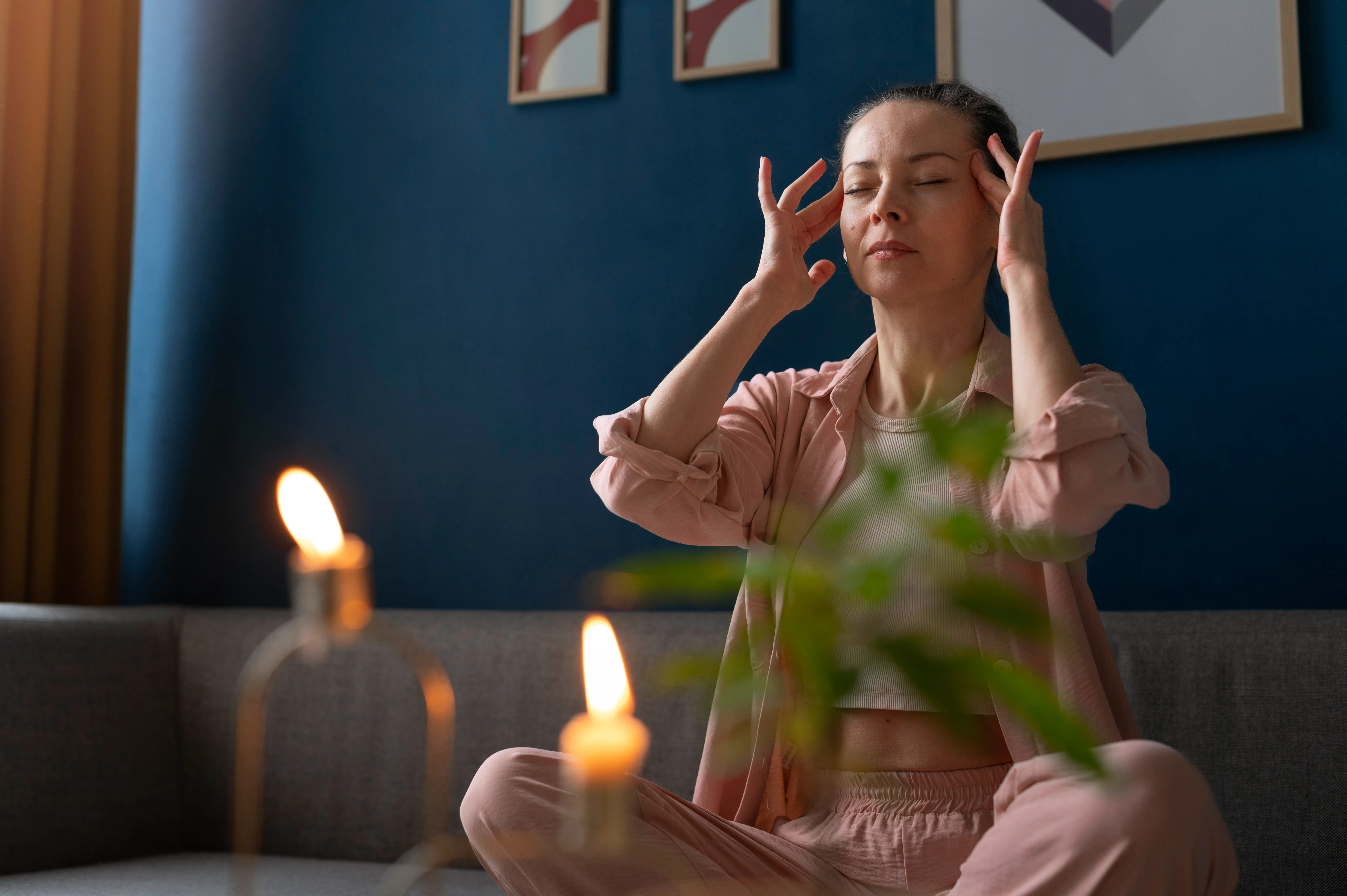A person sitting calmly with eyes closed in a softly lit room, resting fingertips at the temples.